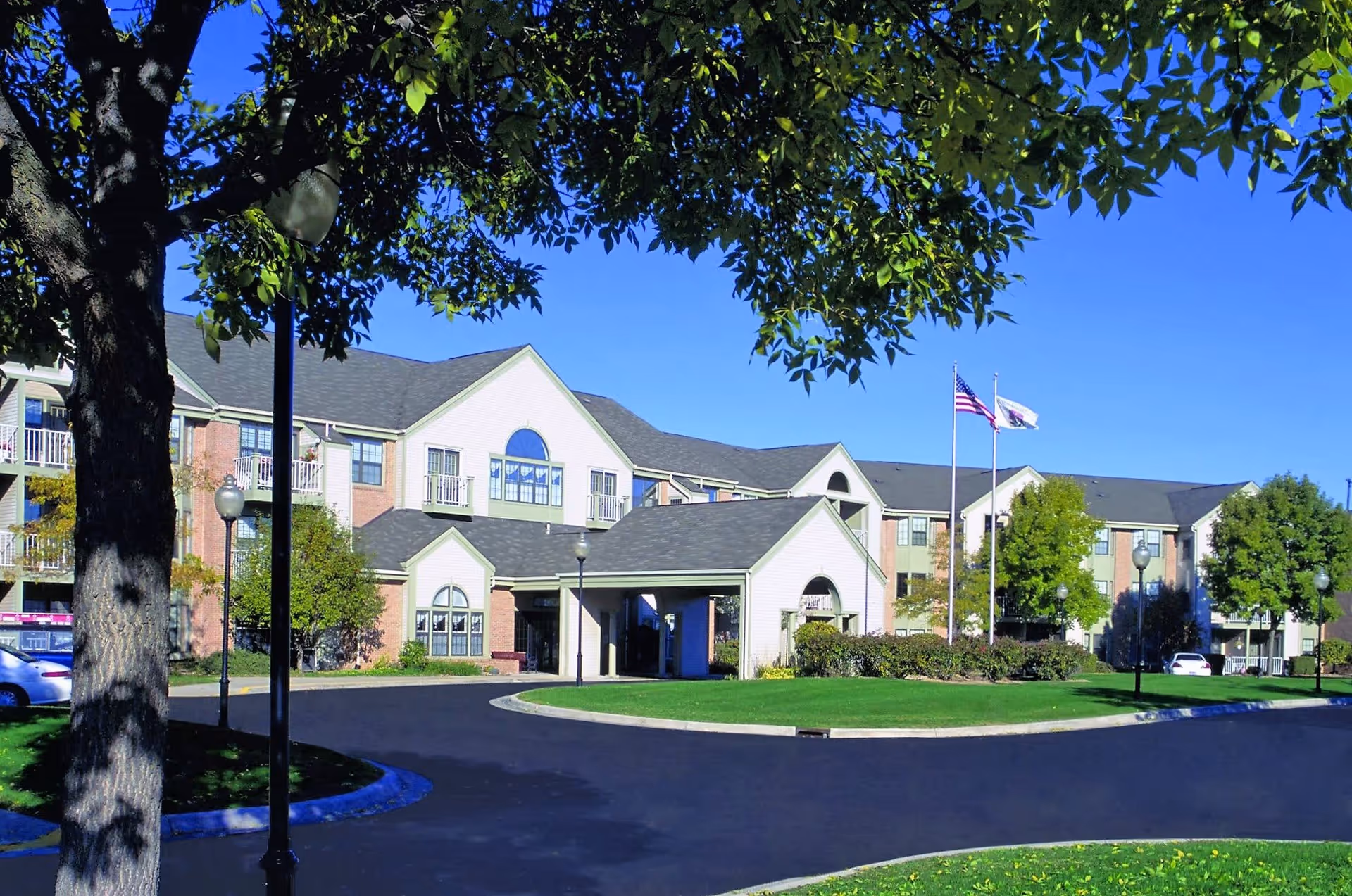 Exterior view of Rosehaven Manor, a multi-story senior living facility with a covered entrance, surrounded by green lawns, trees, and flagpoles displaying the American flag and another flag under a clear blue sky.