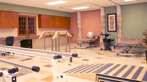 A physical therapy or rehabilitation room with parallel bars for walking exercises, exercise mats, an exercise bike, a large exercise ball, and wooden cabinets along the wall. The room has a tiled floor with a pattern and curtains on the windows.