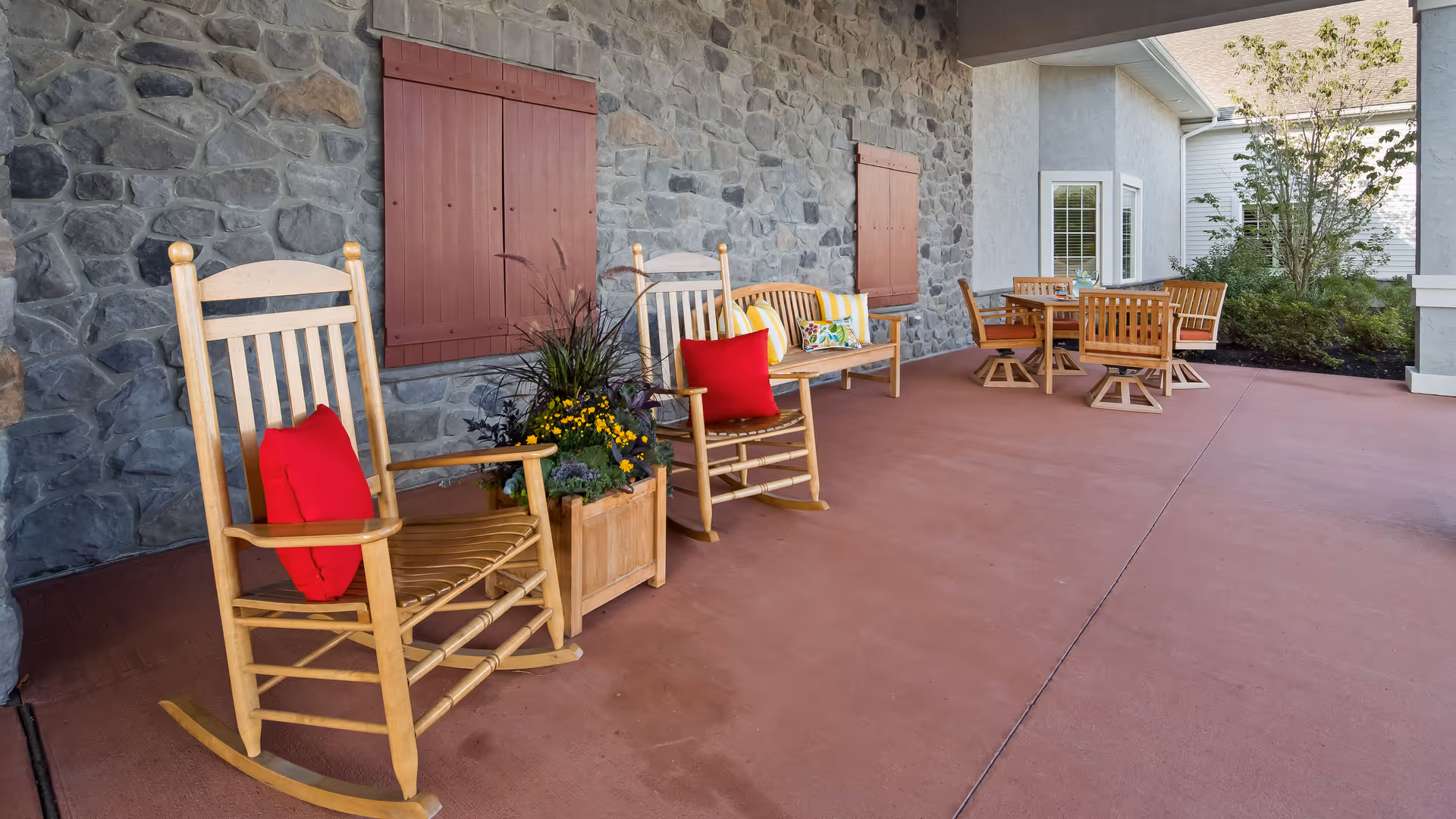 Covered outdoor patio area with two wooden rocking chairs with red cushions, a wooden bench with colorful pillows, a planter with flowers, and a wooden table with four chairs. The patio has a stone wall with closed wooden shutters and a view of greenery and a neighboring building.