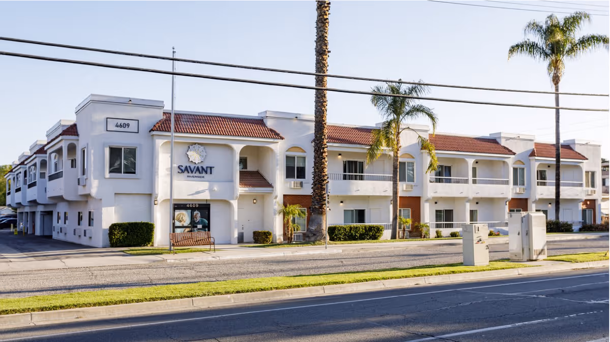 Exterior view of Savant of Riverside, a two-story white building with red-tiled roof, palm trees in front, and a bench near the entrance. The building has multiple windows and balconies, with the address number 4609 visible.