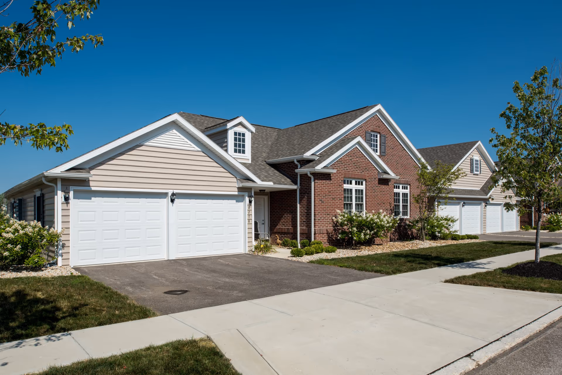 Exterior view of a single-story residential building with a combination of beige siding and red brick walls, featuring multiple white garage doors, windows with shutters, and a well-maintained lawn and sidewalk under a clear blue sky.