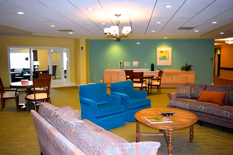 A brightly lit common lounge with sofas, two blue armchairs, a round wooden coffee table and a dining area against a teal accent wall.
