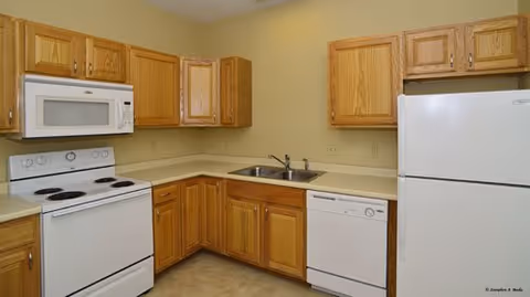 A kitchen with wooden cabinets, a white electric stove with four burners, a white microwave above the stove, a double stainless steel sink, a white dishwasher, and a white refrigerator. The countertops are light-colored and the walls are painted beige.