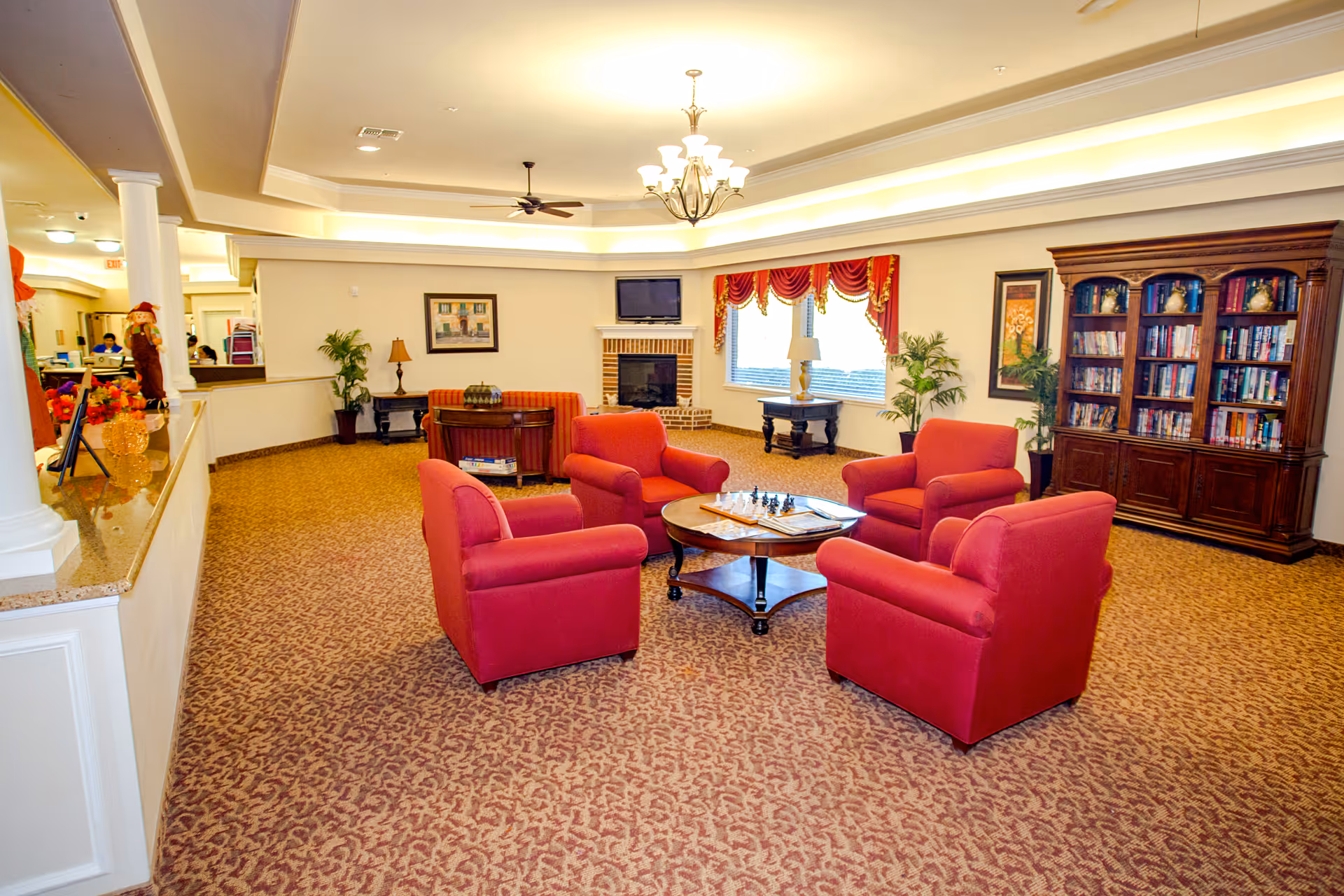 A spacious living room area with four red armchairs arranged around a wooden coffee table with a chessboard on it. The room has patterned carpet, a chandelier, ceiling fan, a fireplace with a TV mounted above it, a large window with red curtains, a wooden bookshelf filled with books, and decorative plants and lamps.