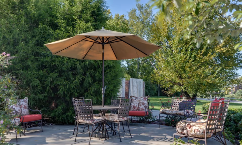 Outdoor patio area with a round table and four chairs under a large beige umbrella, surrounded by additional cushioned chairs with floral and red cushions, set on a stone patio with lush green trees and bushes in the background.