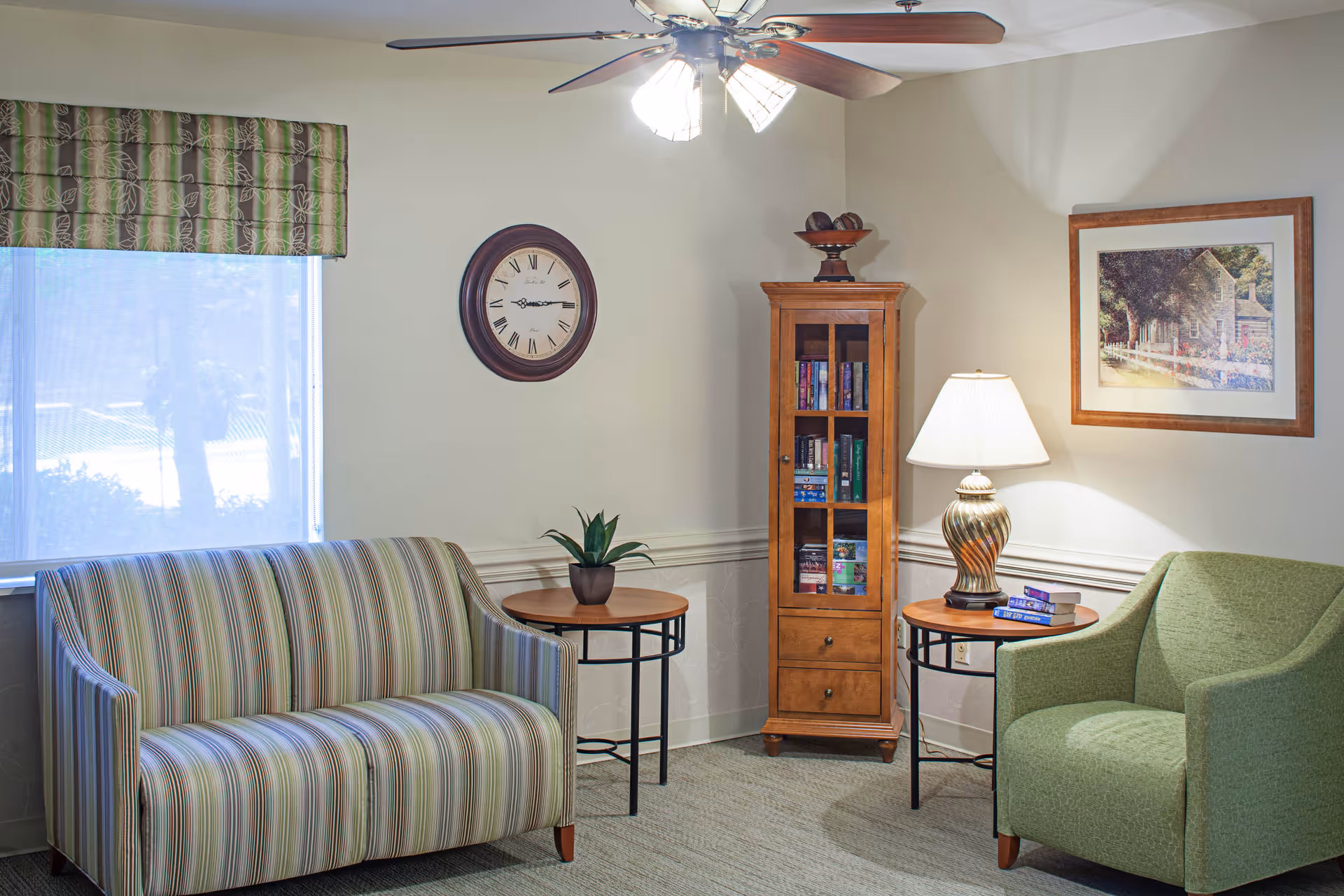 A cozy living room area with a striped sofa on the left, a green armchair on the right, two round wooden side tables with black metal legs, a small potted plant on one table, and a lamp with a decorative base on the other. A tall wooden cabinet with glass doors containing books stands between the tables. A wall clock with Roman numerals hangs above the sofa, and a framed picture of a house and garden is on the wall above the armchair. A ceiling fan with lights is visible on the ceiling.