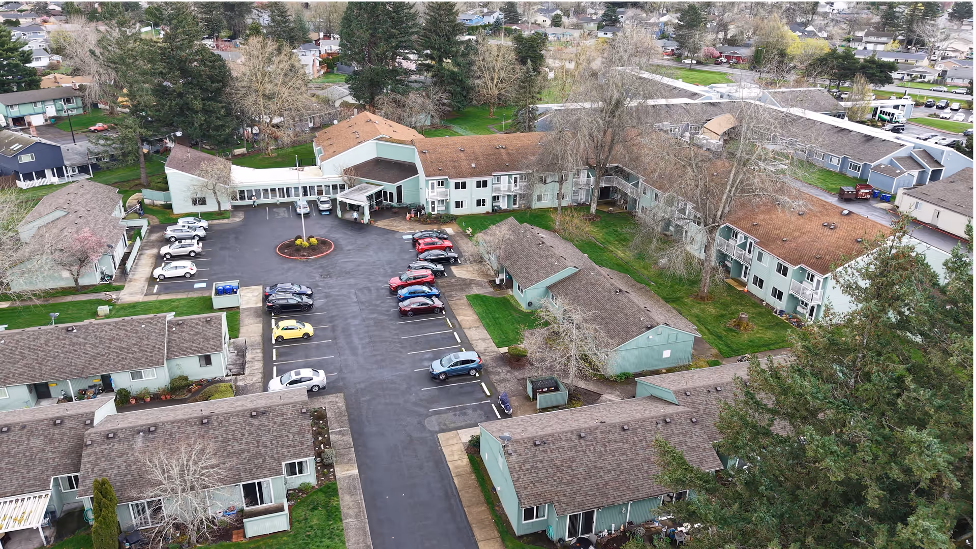 Aerial view of Fairlawn of Olympus Retirement Living facility showing multiple connected buildings with green exterior walls and brown roofs, surrounded by trees and residential houses. There is a parking lot with several cars parked near the main entrance of the facility.