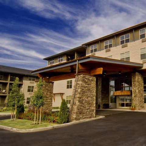 Exterior view of a multi-story senior living facility with stone pillars supporting a covered entrance. The building has multiple windows and is surrounded by small trees and landscaped greenery under a partly cloudy sky.