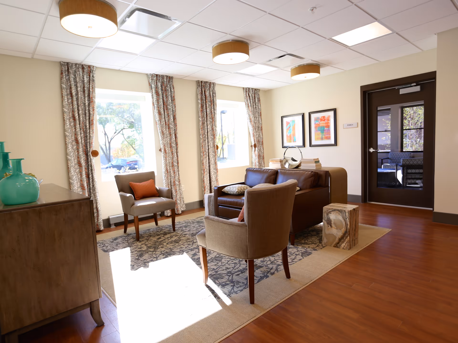 A bright and cozy living room area with two beige armchairs and a brown leather loveseat arranged around a patterned area rug. There are three large windows with patterned curtains letting in natural light. A wooden cabinet with turquoise vases is on the left, and two colorful abstract paintings hang on the wall near a dark brown door labeled 'Porch'. The floor is wood with a beige rug under the seating area.