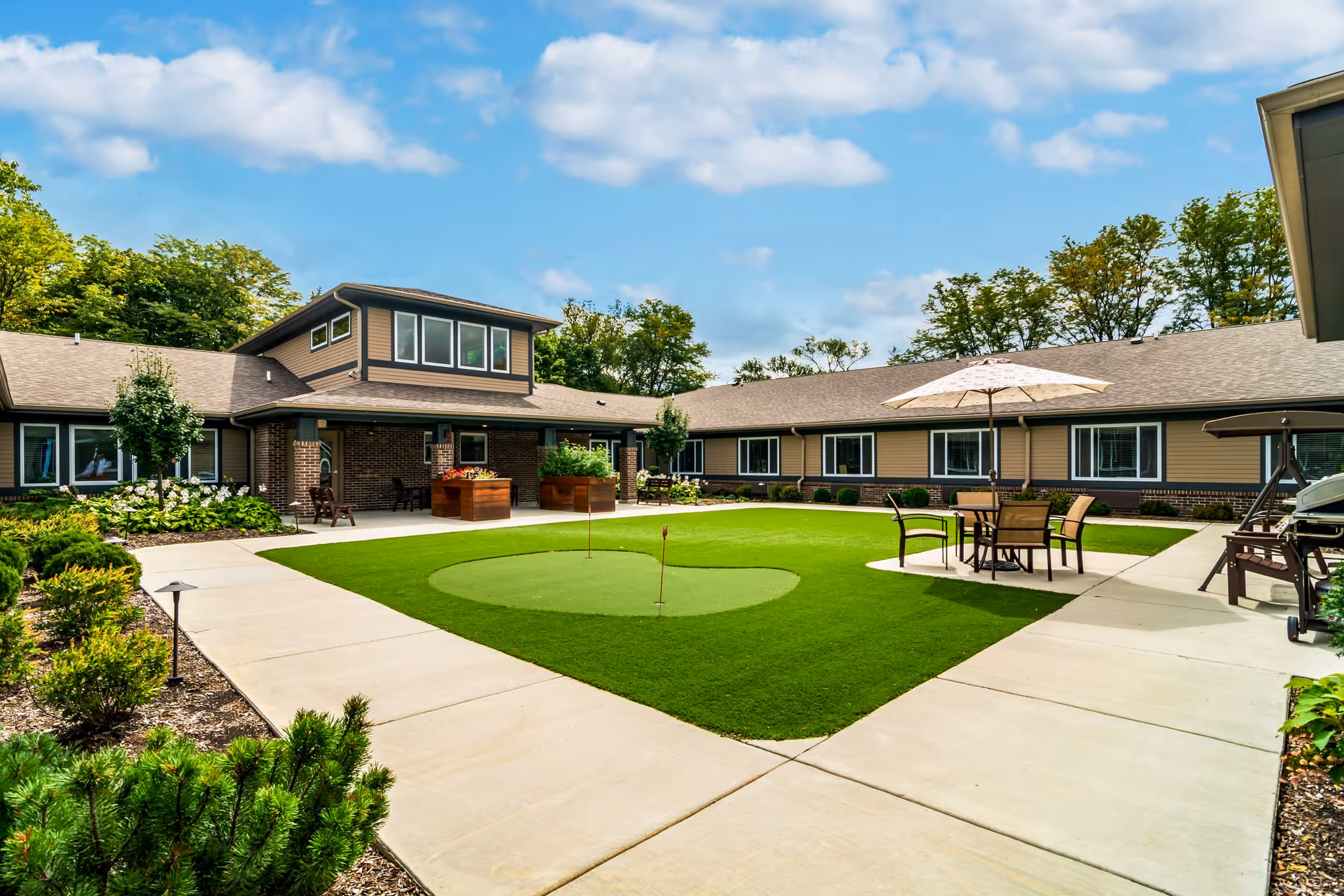Outdoor courtyard area of a senior living facility with a putting green in the center, surrounded by a concrete walkway. There are patio tables with umbrellas and chairs, landscaped greenery, and a single-story building with multiple windows and a covered porch area under a partly cloudy sky.
