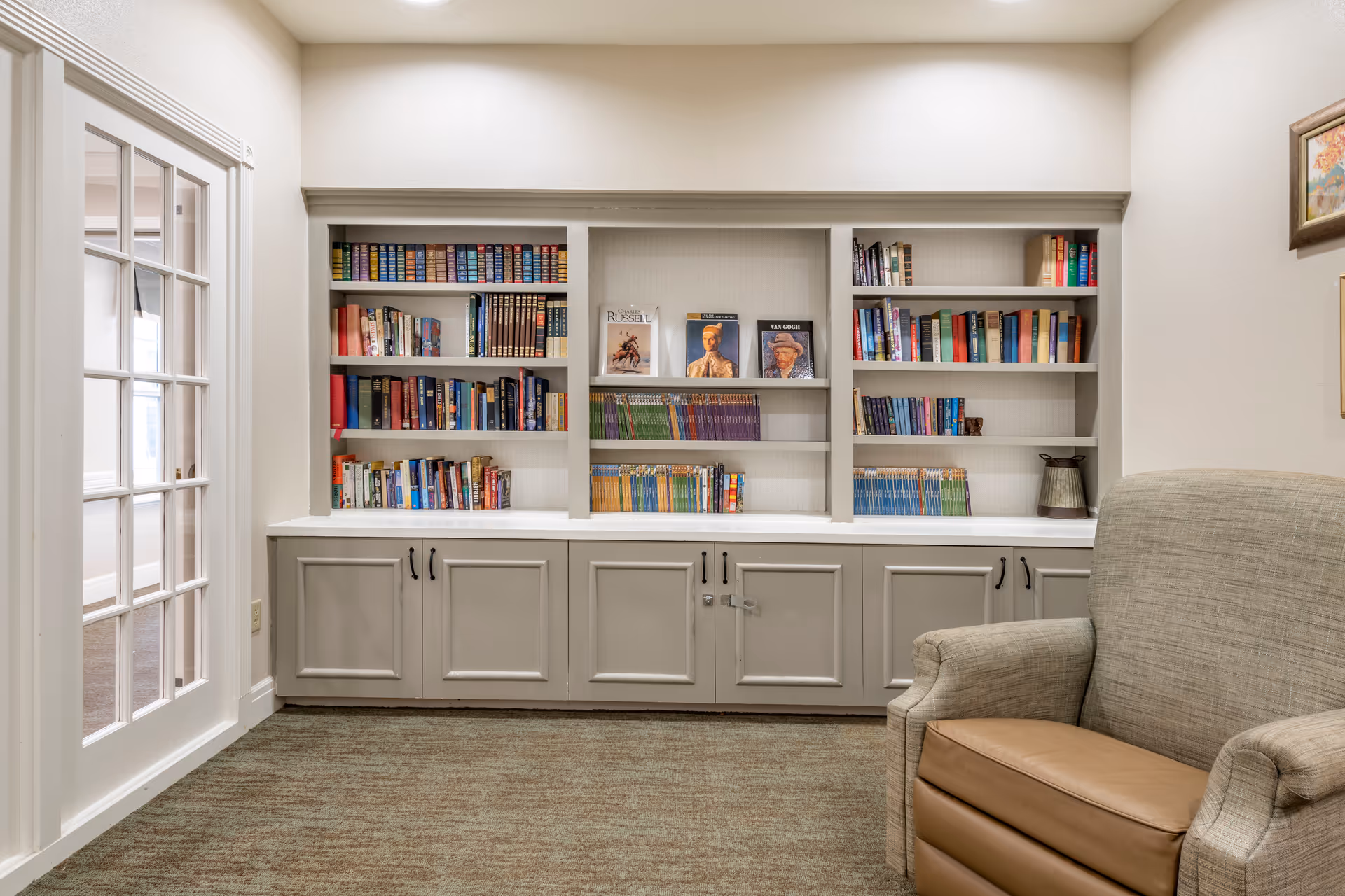 A cozy reading nook in a senior living facility featuring built-in bookshelves filled with books, a comfortable upholstered armchair with a leather seat cushion, and a glass-paneled door on the left side. The room has neutral-colored walls and carpeted flooring.