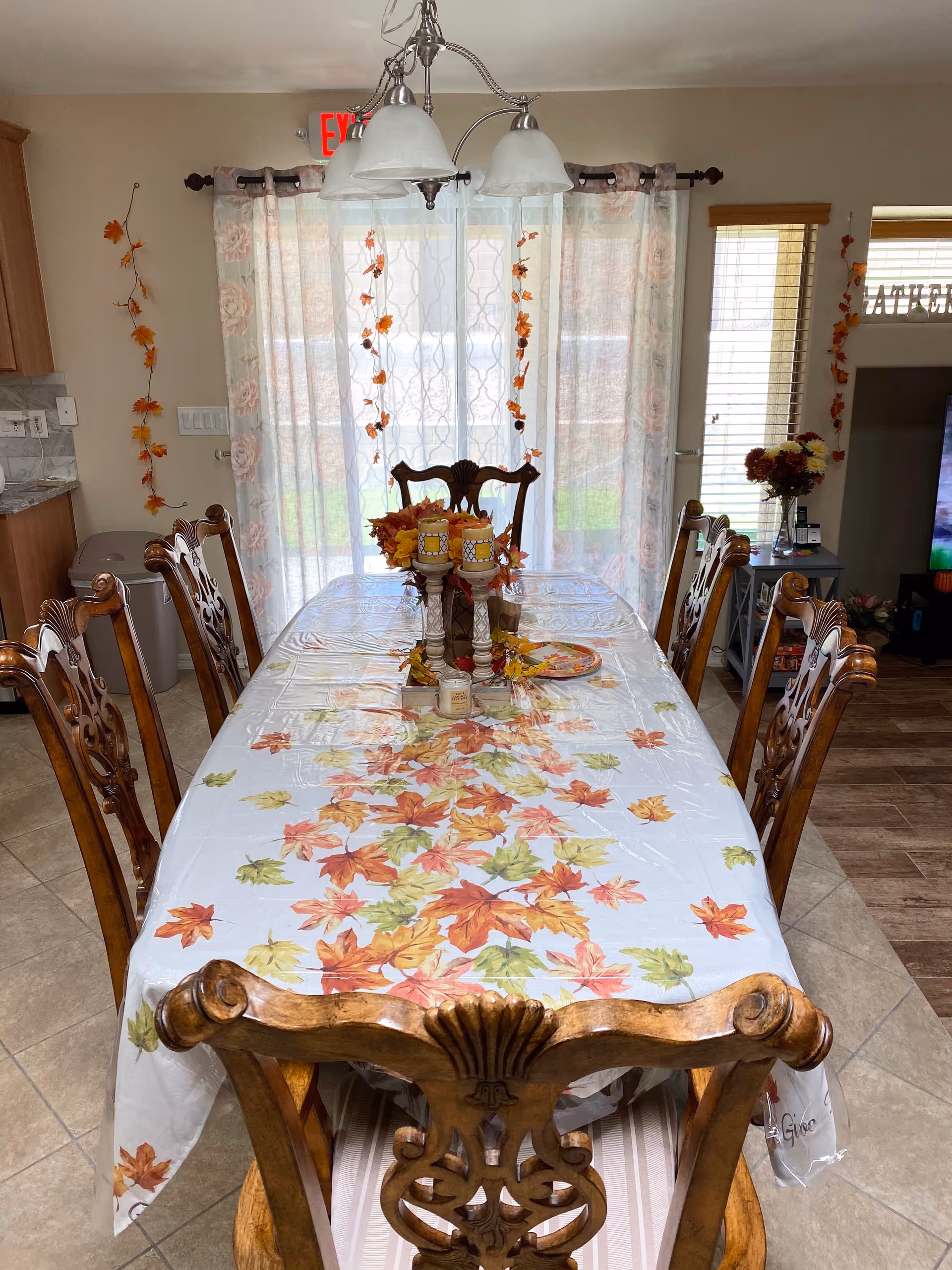 A dining room with a wooden dining table covered with a white tablecloth decorated with autumn leaves. Six ornate wooden chairs surround the table. On the table are three candles on holders and a floral centerpiece. Behind the table are sheer curtains with floral patterns and autumn leaf garlands hanging. To the left is a kitchen counter and a trash bin, and to the right is a small side table with flowers and a TV partially visible.