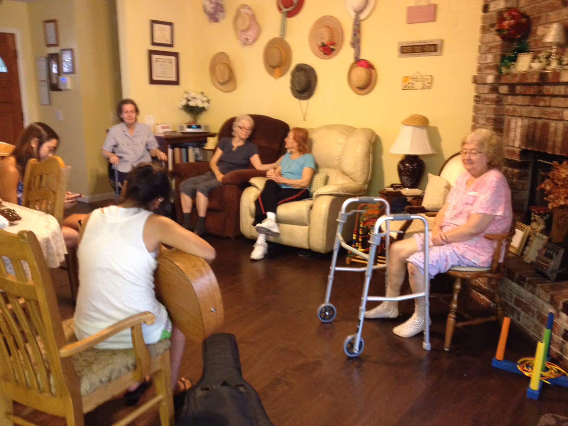 A group of elderly women and younger adults sitting and interacting in a cozy living room with wooden floors, a brick fireplace, and a wall decorated with hats. One elderly woman is seated near a walker, while others are seated on chairs and recliners, engaged in conversation or activities.