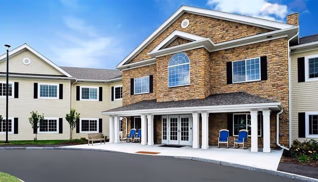 Front exterior view of a two-story senior living facility named The Heron House, featuring a combination of stone and beige siding, multiple windows with black shutters, a covered entrance with white columns, and blue chairs on the porch.