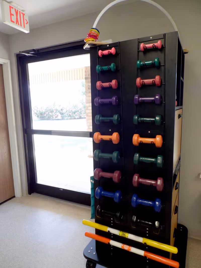 A rack holding various colorful dumbbells and exercise bars is positioned near a glass door with an exit sign above it. The room has light-colored walls and flooring.