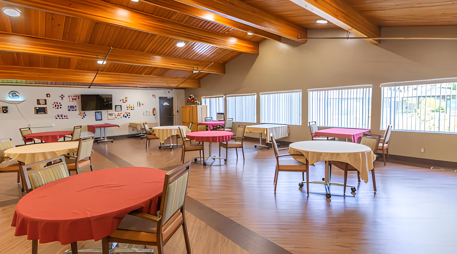 A spacious dining room with multiple round tables covered with red and beige tablecloths, surrounded by chairs. The room has large windows with vertical blinds allowing natural light to enter, and a wooden ceiling with exposed beams. There is a television mounted on the far wall and some decorations and furniture along the walls.