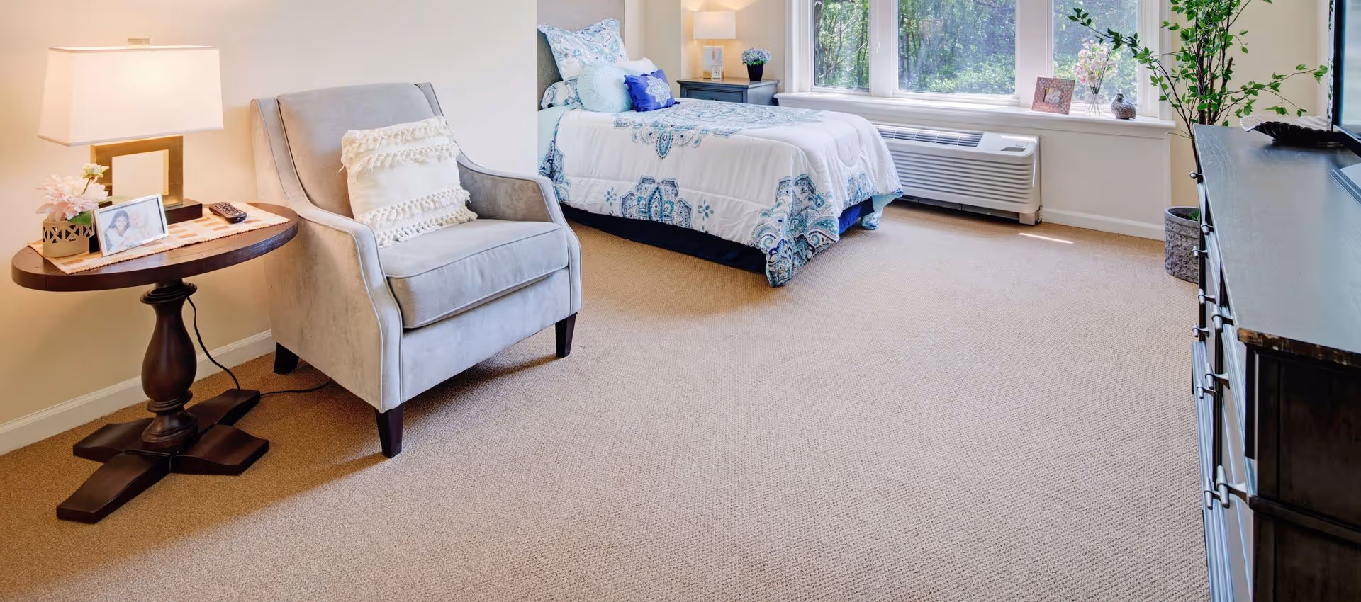 A bright furnished bedroom with an upholstered armchair and side table, a bed by large windows, and a dresser along the wall.