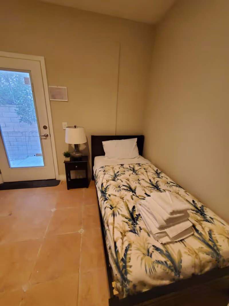 A small bedroom with a single bed covered with a floral patterned blanket and a stack of folded white towels on top. Next to the bed is a dark wooden nightstand with a lamp and a small plant. There is a glass door with blinds leading outside, and the floor is tiled.