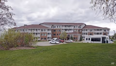 A large senior living facility exterior with a bus and parked cars in front.
