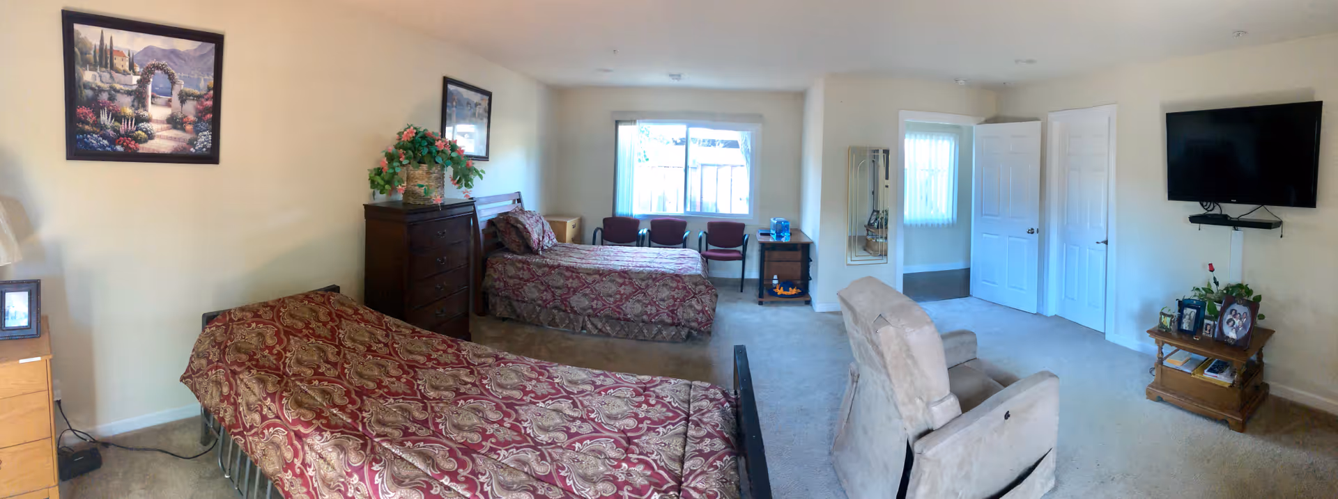 A spacious bedroom with two twin beds covered in patterned red and gold bedspreads. The room features a wooden dresser with a plant on top, three chairs near a window with vertical blinds, a beige recliner chair, a wall-mounted flat-screen TV, and a small wooden table with framed photos and a plant. The walls are light-colored and decorated with framed artwork.