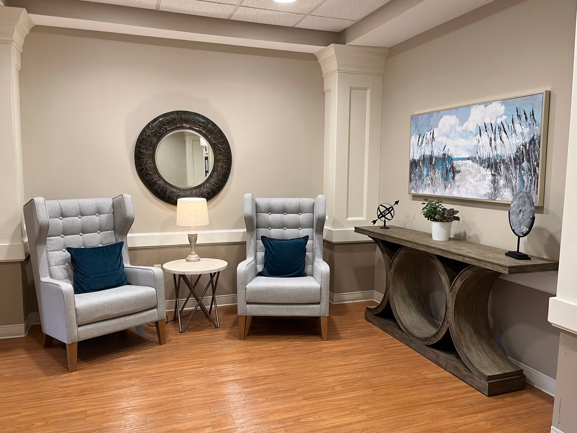 A cozy seating area with two gray wingback chairs, a small round table and lamp between them, a decorative round mirror, and a console table with artwork on the wall.