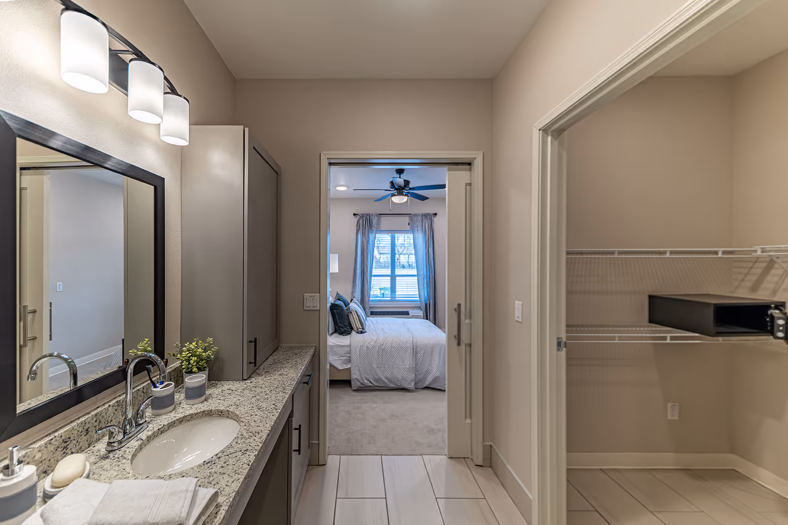 View of a bathroom vanity with a granite countertop, sink, and mirror on the left side. The bathroom has light-colored walls and tile flooring. Through an open doorway, a bedroom with a bed, pillows, curtains, and a ceiling fan is visible. On the right side, there is an open closet with wire shelving and a black safe.