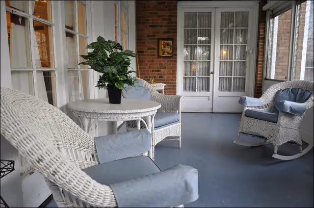 A cozy enclosed porch area with white wicker furniture including a round table and three chairs with light blue cushions. A potted green plant is placed on the table. The porch has large windows and white French doors with lace curtains, and a brick wall with a small framed picture.