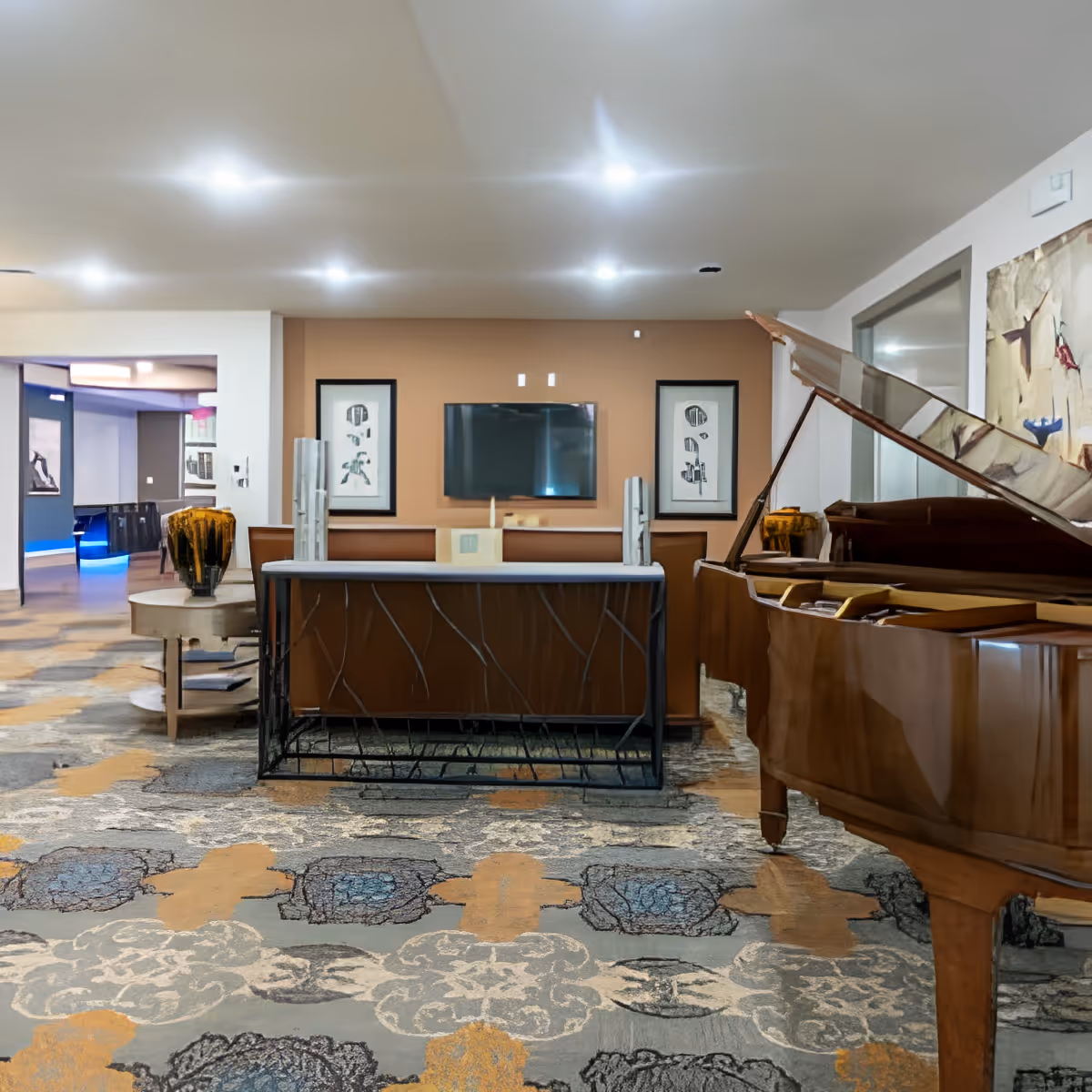 Interior view of a common area in a senior living facility featuring a grand piano on the right, a patterned carpet, a console table with decorative items, a wall-mounted flat screen TV, and framed artwork on a beige accent wall.