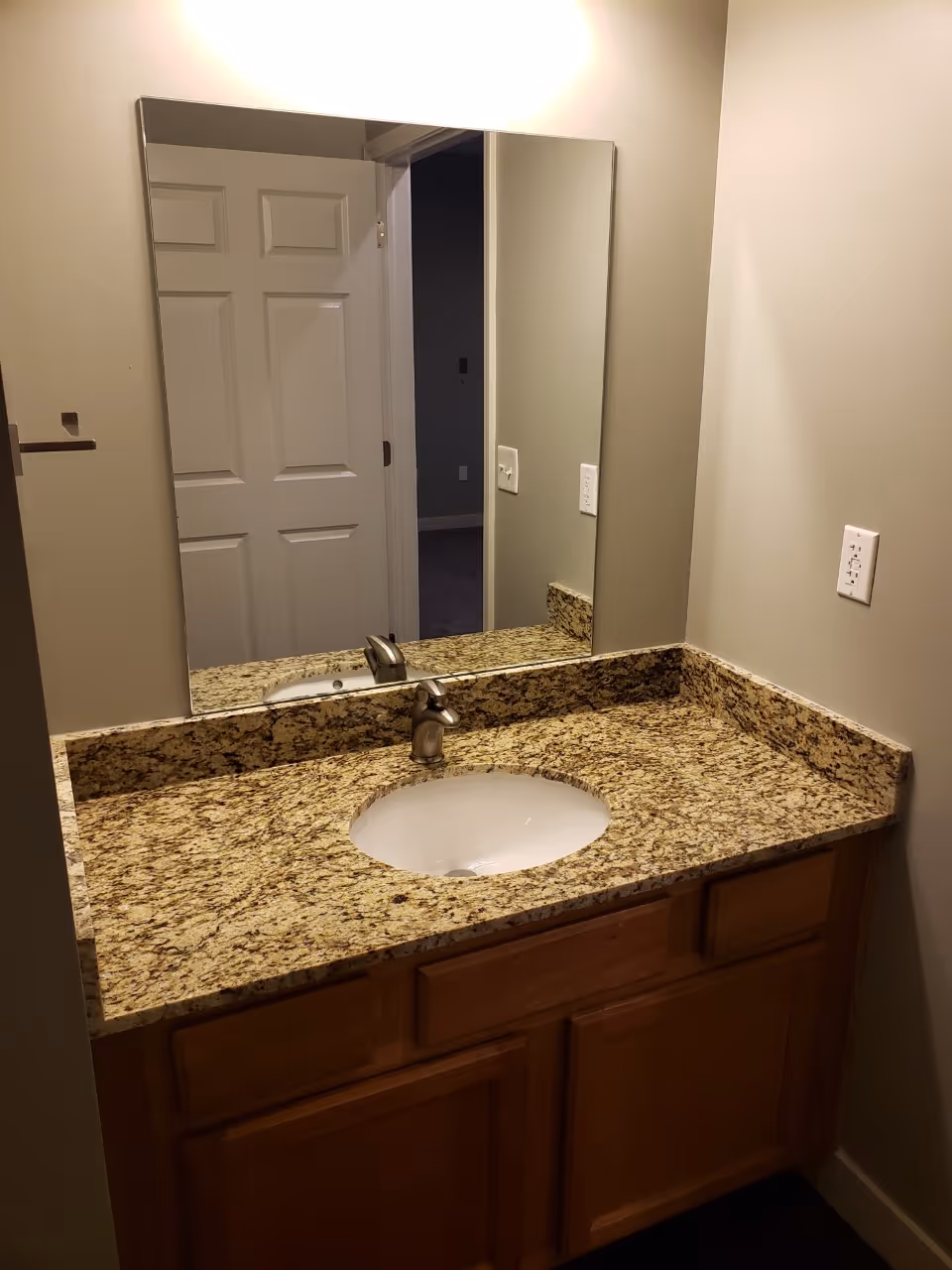 Bathroom vanity with a granite countertop, an undermount sink, and a single-handle faucet. A large rectangular mirror is mounted on the wall above the sink, reflecting a white paneled door and part of an adjacent room. The walls are painted beige, and there is a white electrical outlet on the right wall.
