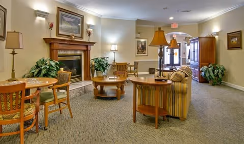 A cozy living room area in Ashland Villa featuring a fireplace with a wooden mantel, framed artwork above it, several potted plants, a striped sofa, wooden tables with lamps, and multiple chairs arranged around small round tables. The room has soft lighting and a carpeted floor, with an exit door visible in the background.