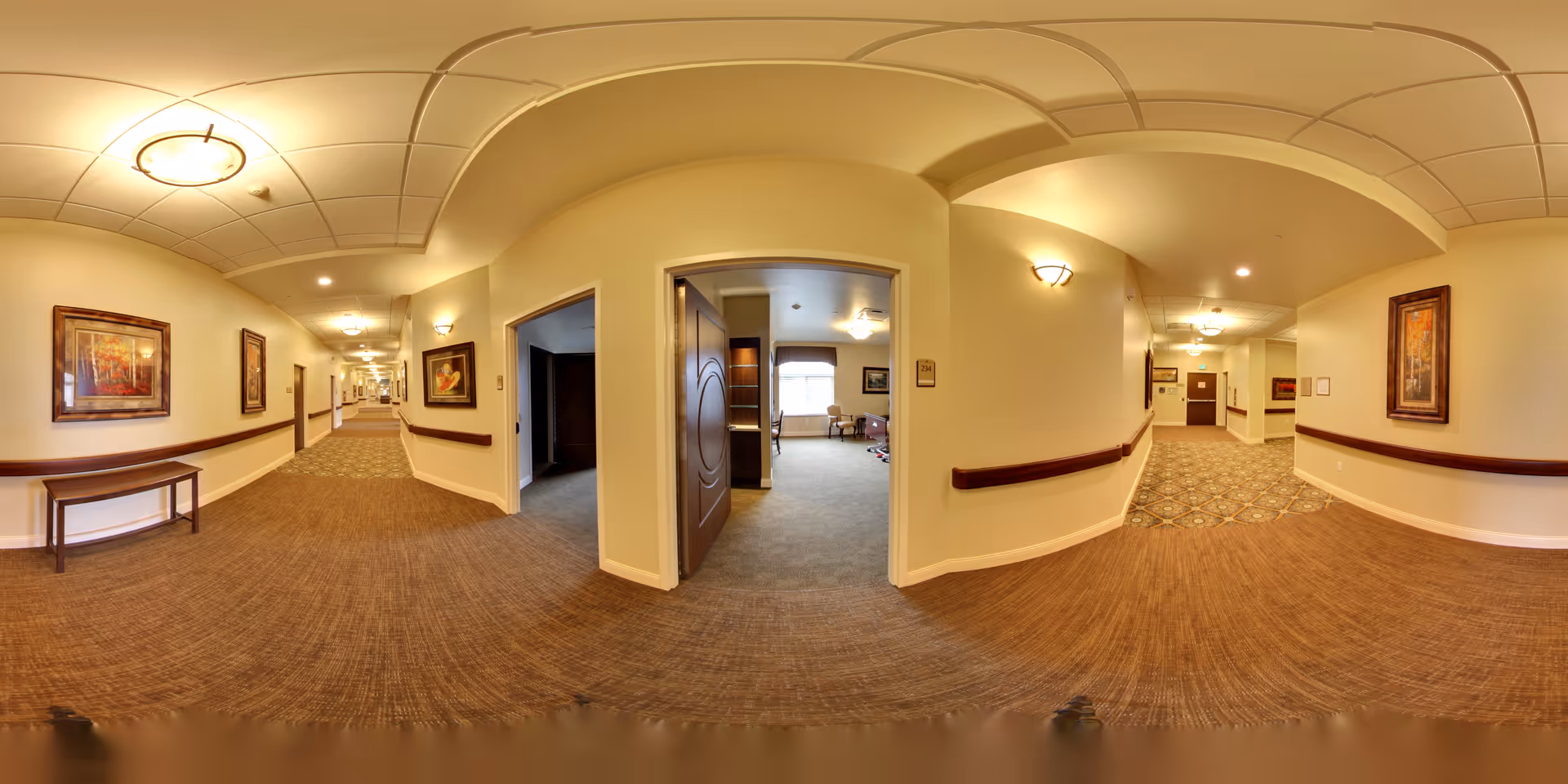 A wide-angle view of a well-lit hallway in a senior living facility with beige walls, carpeted floors, and several framed paintings on the walls. The hallway has handrails on both sides and leads to multiple rooms, one of which is open showing a sitting area with chairs and a window letting in natural light.