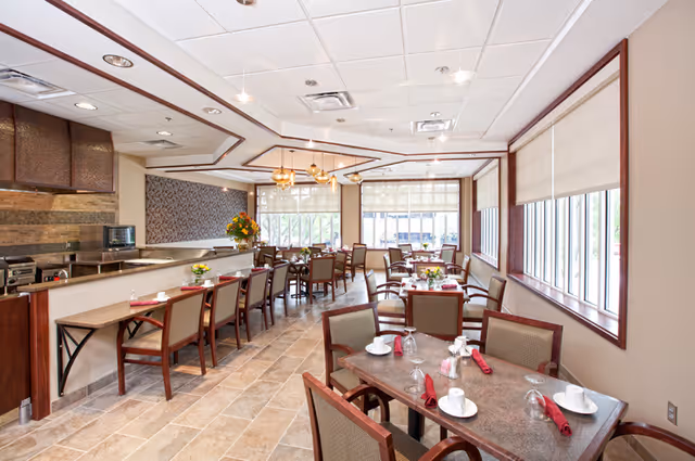A bright dining room with multiple tables and chairs arranged neatly. Each table is set with white cups, glasses, and red napkins. Large windows with blinds allow natural light to fill the room. There is a counter area with additional seating and a kitchen area visible in the background.