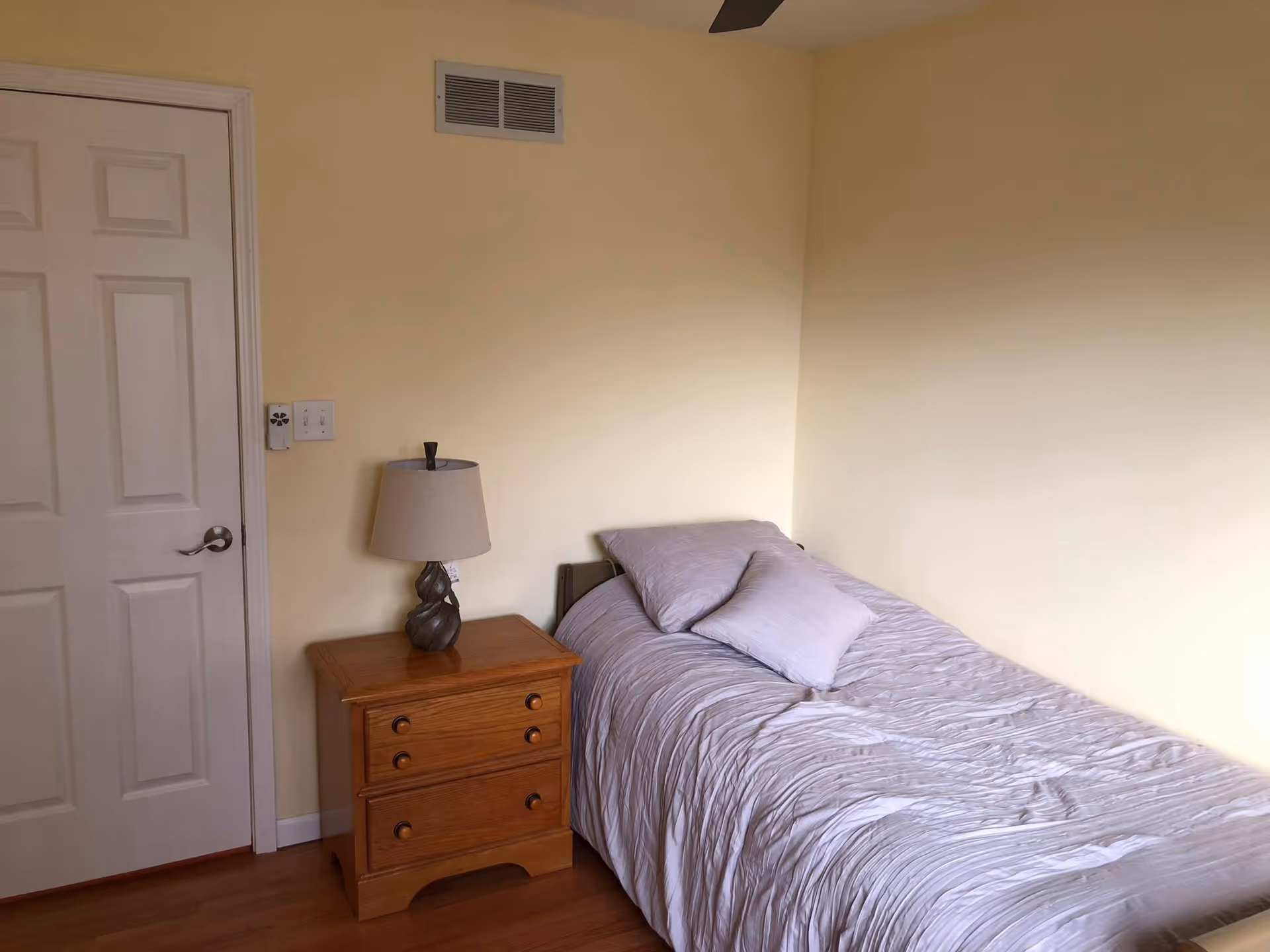 A small bedroom with a single bed covered in light gray bedding and two pillows. Next to the bed is a wooden nightstand with three drawers and a decorative lamp on top. The walls are painted light yellow, and there is a white door with a silver handle on the left side of the image.