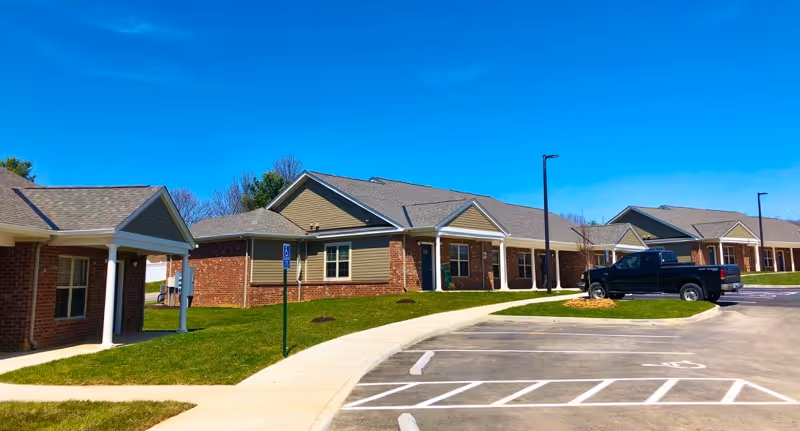 Exterior view of single-story brick and siding apartment buildings with a parking lot in front under a clear blue sky. There is a black pickup truck parked in the lot and a sidewalk running alongside the buildings.