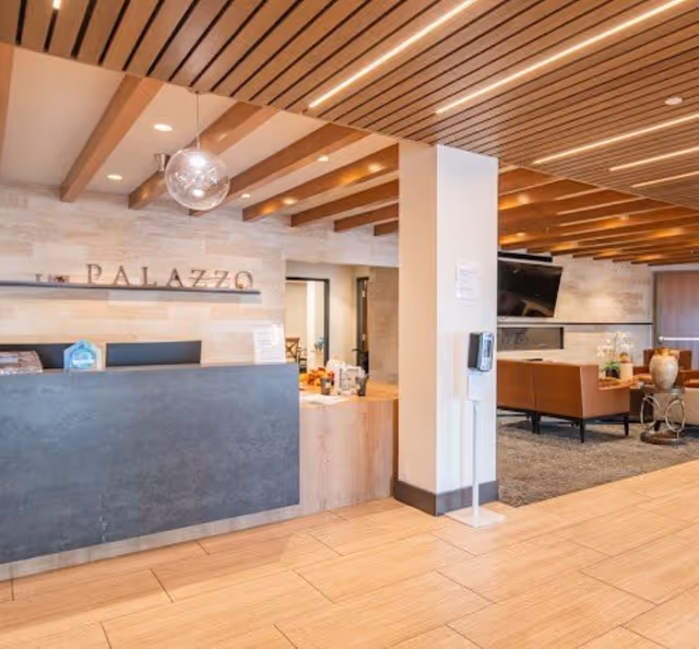 Reception area of The Palazzo Skilled Nursing & Rehab featuring a modern front desk with a dark stone facade, wooden ceiling beams, and a seating area with brown leather chairs and a wall-mounted TV in the background.