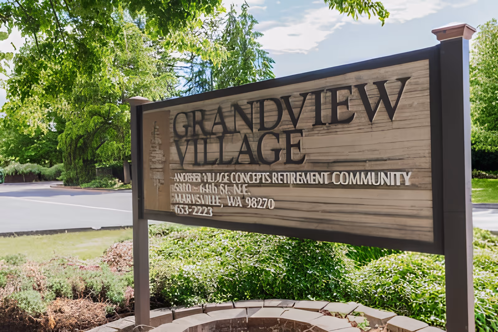 A wooden sign for Grandview Village, a retirement community by Village Concepts, standing outdoors surrounded by green trees and bushes under a partly cloudy sky.