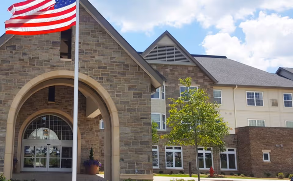 Exterior view of a retirement community building with stone and brick facade, large arched entrance, multiple windows, a small tree in front, and an American flag on a flagpole waving in the breeze under a partly cloudy sky.