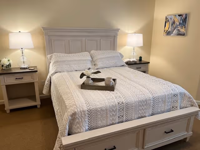 A neatly made bed with a white patterned quilt and two pillows in a bedroom. The bed has a wooden headboard and footboard painted in a light color. There are two matching nightstands on either side of the bed, each with a lamp and decorative items. A small tray with a flower and a cup is placed on the bed. A piece of abstract artwork hangs on the wall to the right.