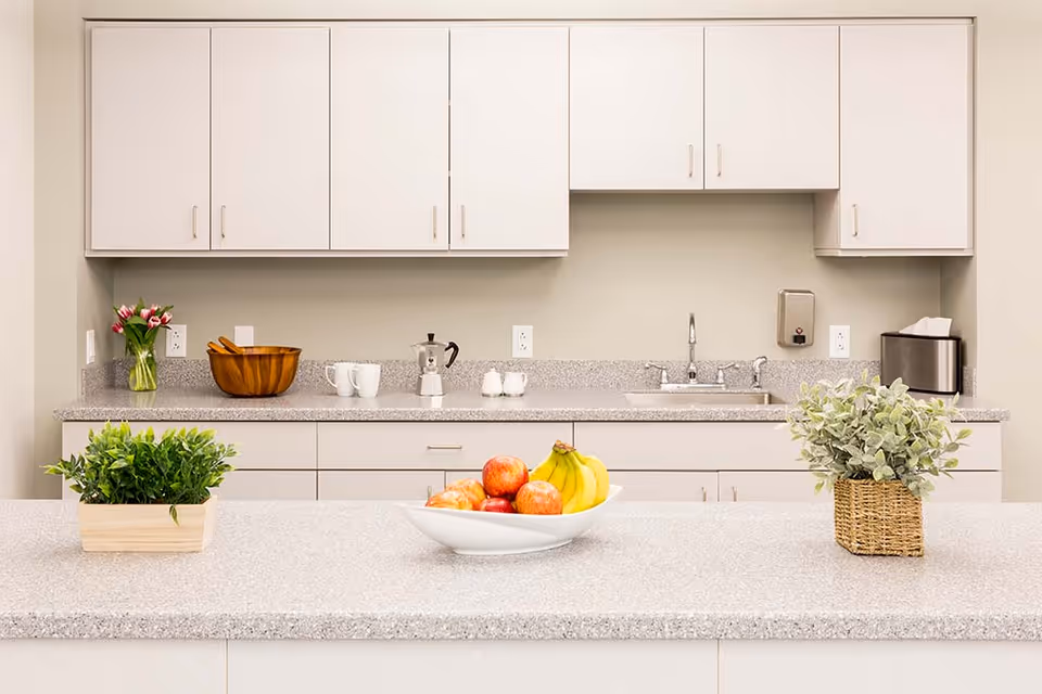 A clean and modern kitchen area with white cabinets and a light gray countertop. On the countertop, there is a white bowl filled with apples and bananas, a small green potted plant in a wooden container, and another green plant in a woven basket. In the background, there is a sink with a faucet, a wooden bowl, two white mugs, a coffee maker, a soap dispenser, a tissue box, and a vase with pink tulips.