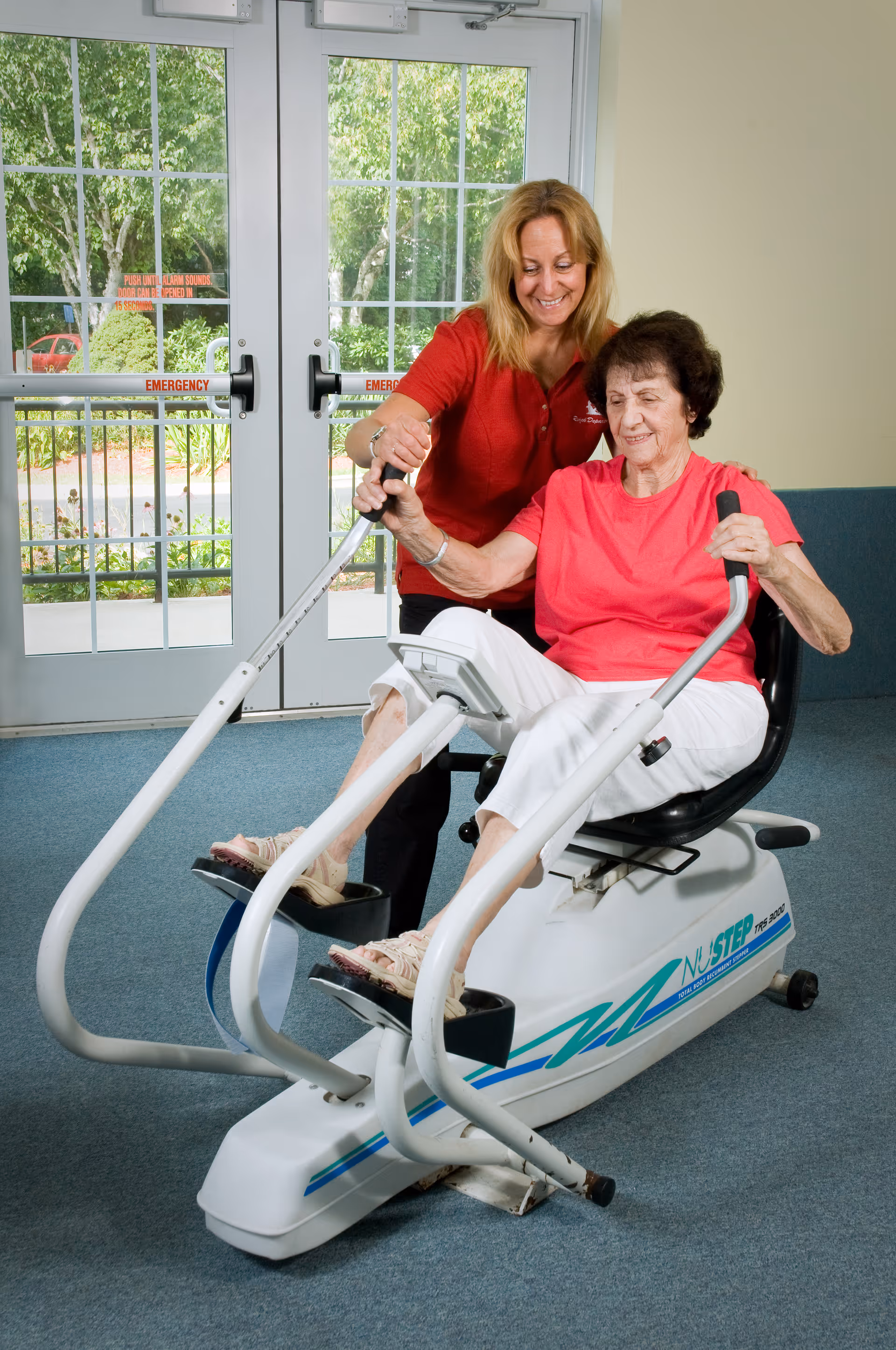 An elderly woman in a pink shirt and white pants is using a NuStep exercise machine indoors, assisted by a smiling caregiver in a red shirt. They are in a room with large glass doors showing greenery outside.