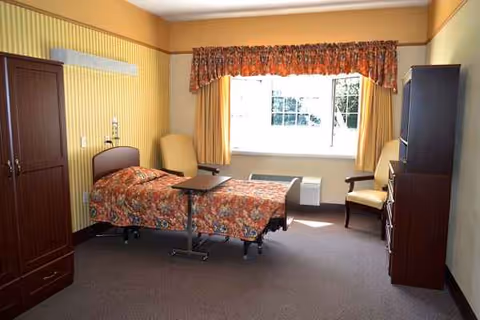 Hospital-style patient room with a single bed, chair, wardrobe, and window with curtains.