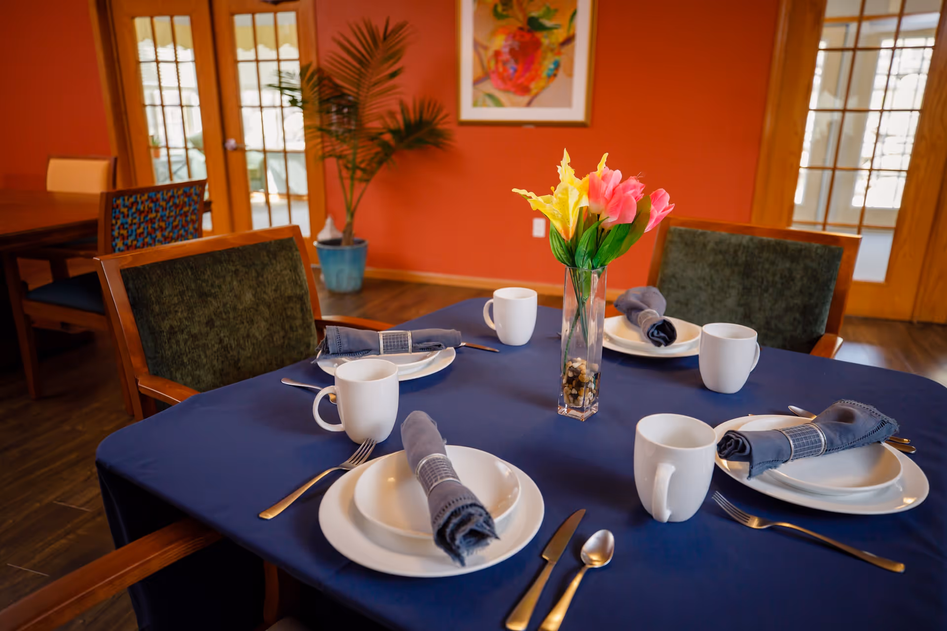 A dining table set for four with white plates, cups, silverware, and blue cloth napkins rolled with napkin rings. A vase with yellow and pink flowers is centered on the table, which is covered with a dark blue tablecloth. The room has wooden floors, orange walls, wooden framed glass doors, a potted plant, and a colorful framed artwork on the wall.