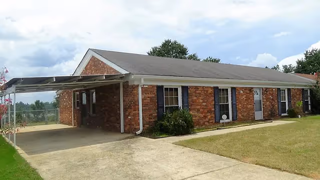 Single-story brick building with a sloped roof, multiple windows with dark shutters, a covered carport on the left side, and a concrete driveway leading up to the entrance. The building is surrounded by a grassy lawn and some bushes, under a partly cloudy sky.