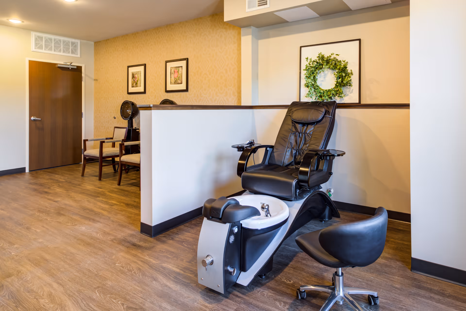 Interior view of a senior living facility salon area with a black leather salon chair and foot basin for hair washing, a black swivel stool, two wooden chairs with beige cushions, framed floral artwork on the walls, and a decorative green wreath on a framed picture.