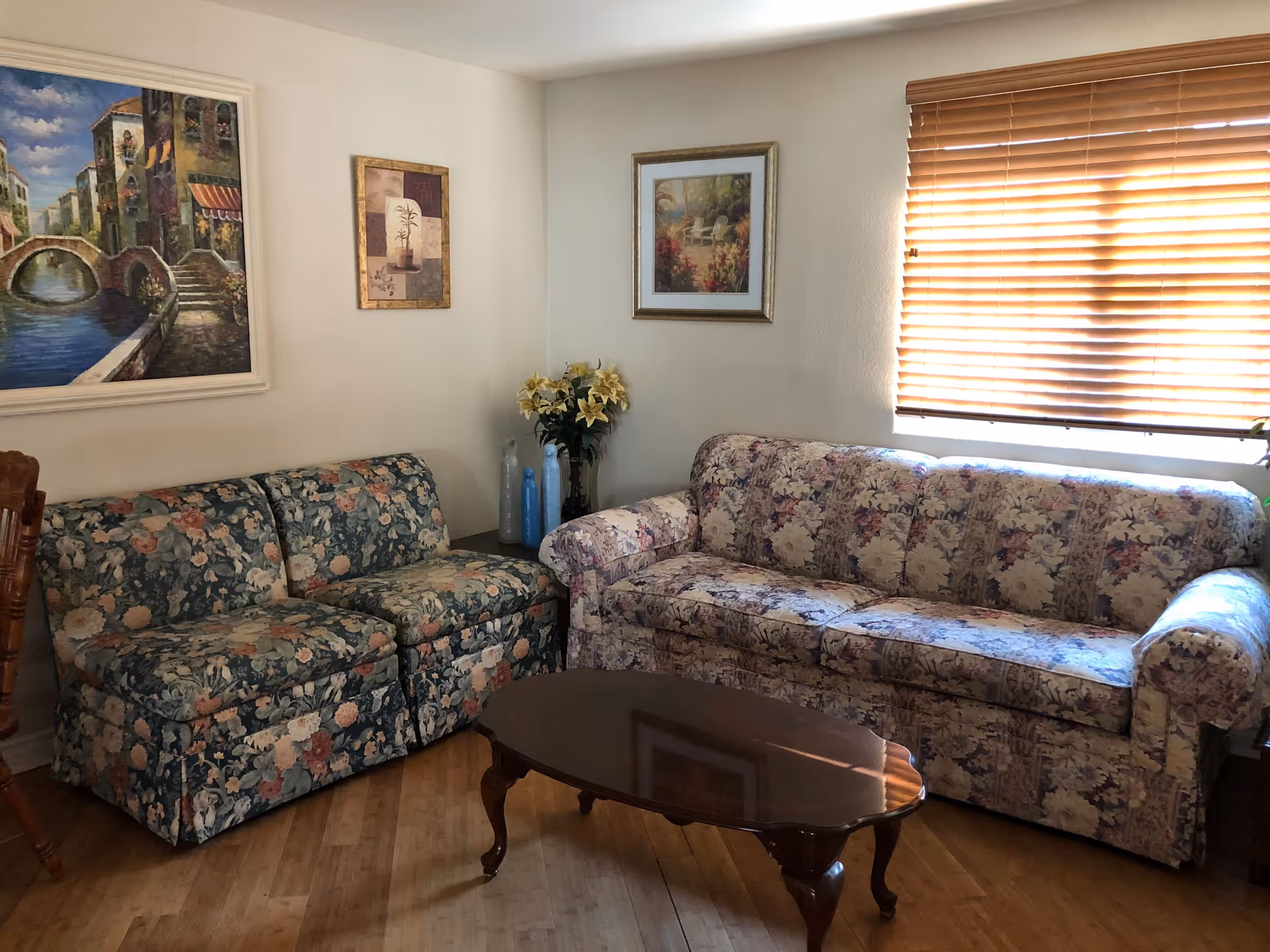 A cozy living room with two floral-patterned sofas, a wooden coffee table, and three framed paintings on the walls. A window with wooden blinds lets in natural light, and a vase with yellow flowers sits on a side table between the sofas.