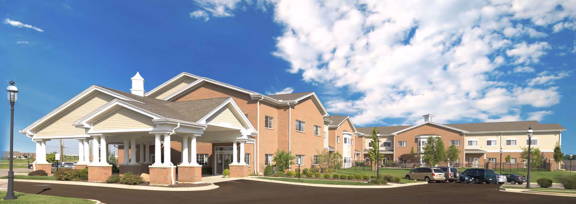 Exterior view of a large two-story senior living facility building with a covered entrance supported by white columns, a parking lot with several cars, green lawns, and a partly cloudy blue sky.