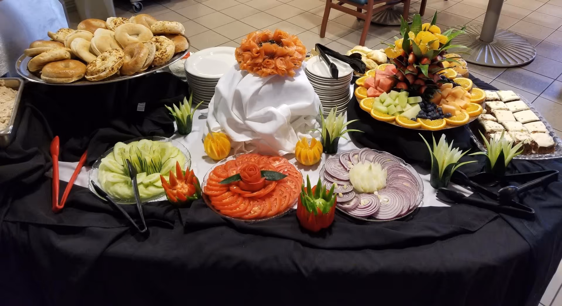 A buffet table with a variety of food items including bagels, sliced tomatoes, cucumbers, onions, a platter of assorted fresh fruit arranged around a pineapple centerpiece, and a plate of dessert bars. The table is covered with a black and white tablecloth and has plates and utensils arranged for serving.