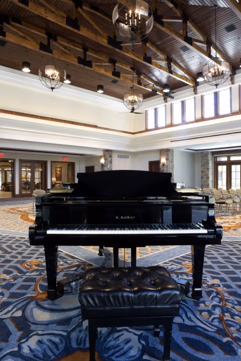 A black K. Kawai grand piano with a matching padded bench in a spacious room featuring a high wooden beamed ceiling with chandeliers, patterned carpet, and large windows letting in natural light.