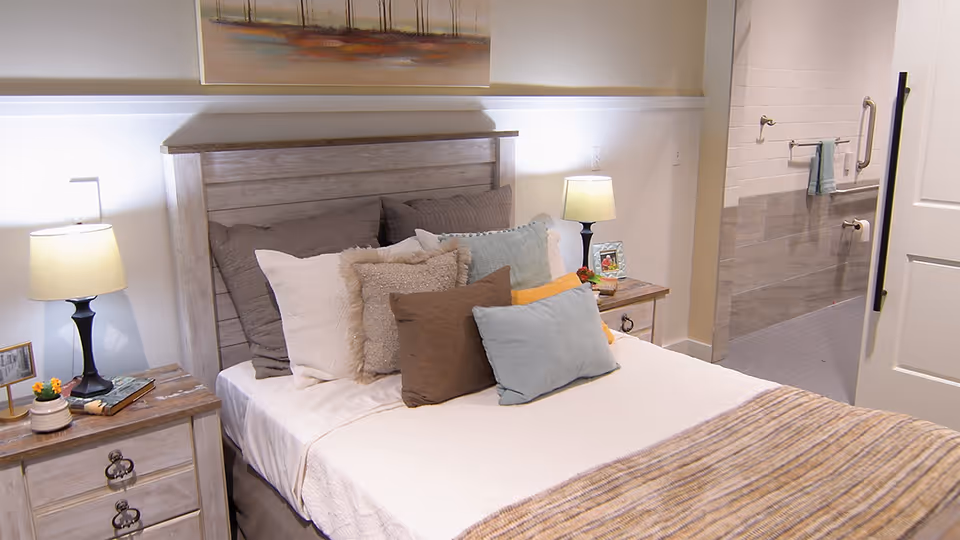 A cozy bedroom with a wooden headboard bed adorned with multiple pillows in neutral and pastel colors. There are two bedside tables with lamps, a small plant, and picture frames. The room has soft lighting and a painting above the bed. An open door reveals a bathroom with grab bars and a towel hanging on the wall.