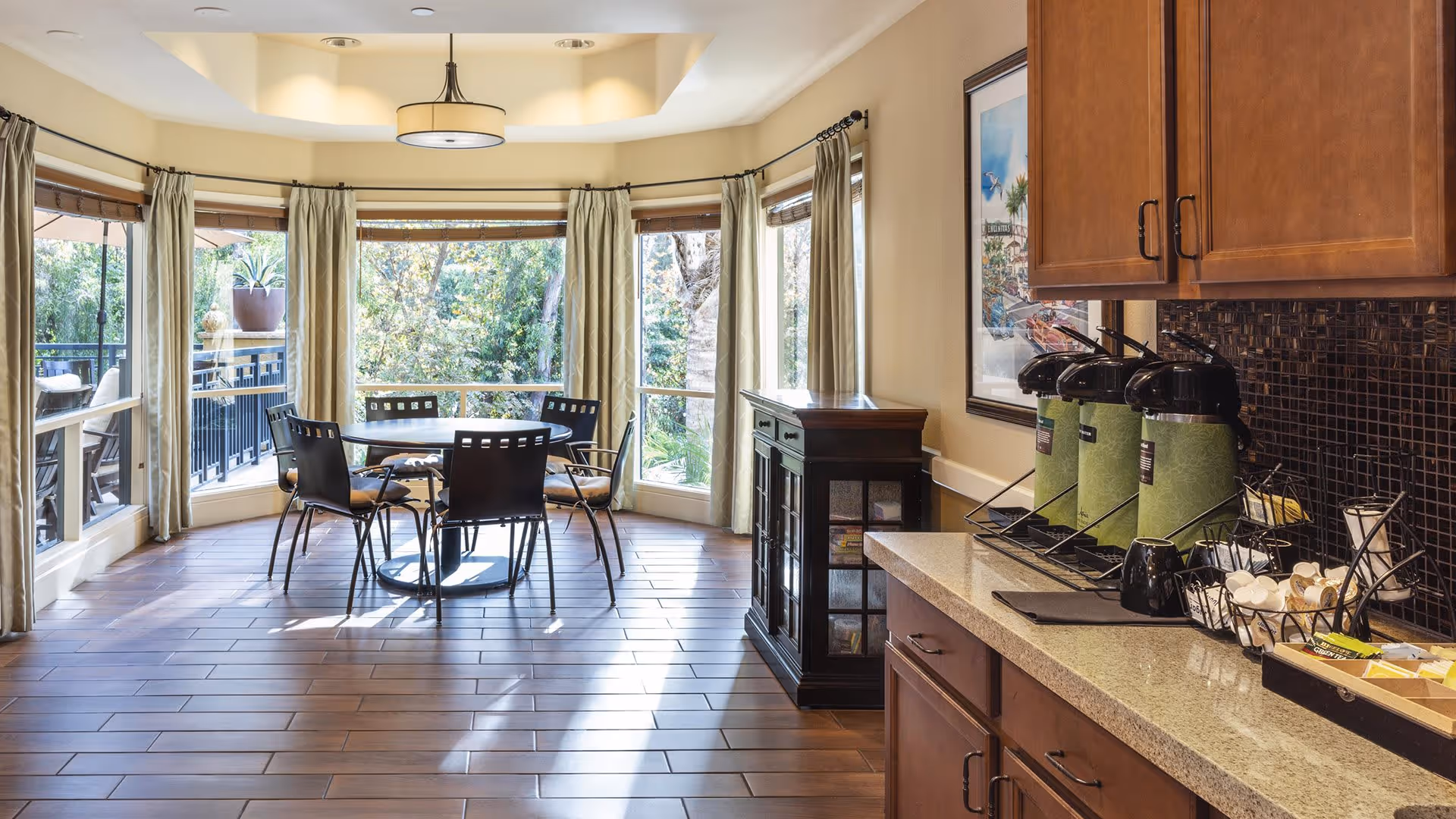 A bright dining area with a round table and four chairs near large windows with curtains, overlooking greenery outside. To the right, there is a countertop with coffee dispensers, cups, and condiments, and wooden cabinets above and below the counter.